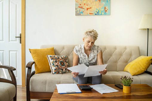 A woman is looking at some documents in her hands while sitting in her living room, worrying about her spending and income