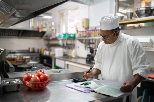 A chef calculating the kitchen expenses An Indian chef is writing up his daily accounts, working on the kitchen finances while standing at a counter in a commercial kitchen energy bill photos stock pictures, royalty-free photos & images