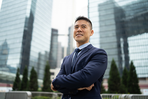 Portrait of a businessman smiling as he looks into the distance, standing with his arms crossed against a background of tall glass covered office buildings in a city