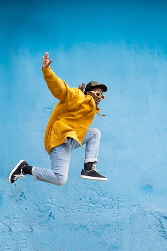 An African man leaping high in the air, with legs akimbo and arms stretched out, wearing modern clothing, baseball cap and sunglasses against a blue wall
