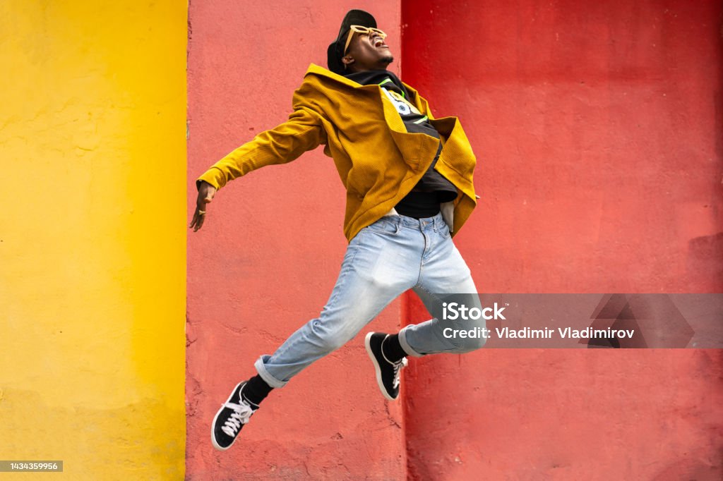 Male urban dancer in the air Young African-American man is mid pose in the air with arms and legs kinked backwards, wearing modern clothing against orange and yellow background Dancing Stock Photo
