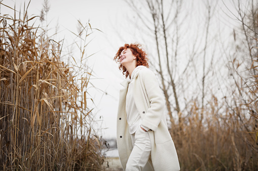 Beautiful young woman in trendy white clothes poses in motion and laughing against the backdrop of an autumn landscape with dry reeds