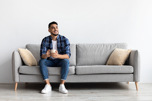 Relax Concept. Happy Arab man drinking coffee sitting on comfortable couch at home in living room and dreaming. Cheerful casual male resting on sofa, enjoying weekend free time, full body length