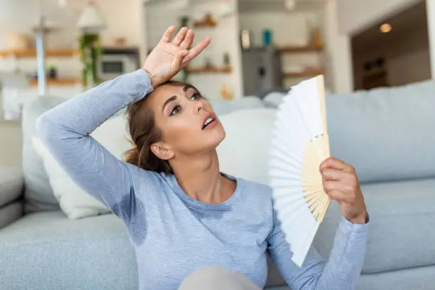 Woman puts head on sofa cushions feels sluggish due unbearable heat, waves hand fan cool herself, hot summer flat without air-conditioner climate control system concept