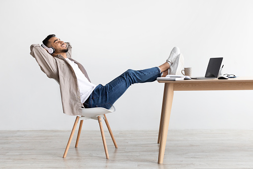 Taking Break From Work. Happy Arabic man with closed eyes sitting on chair, listening to music or podcast in wireless headset, leaning back holding hands behind head sitting with legs on table