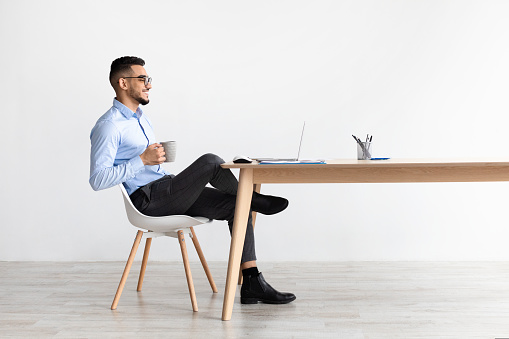 People And Technology. Profile portrait of young smiling Arab man using pc sitting at desk, holding cup of hot coffee. Cheerful guy in glasses watching video, free copy space, full body length