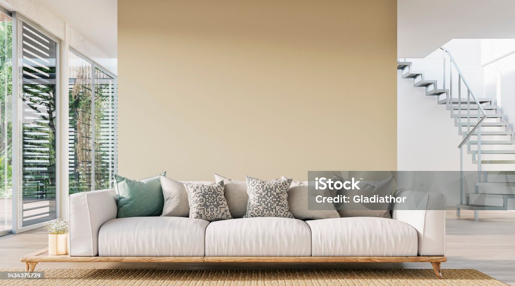 Cozy, luxury, and modern living room with sofa, windows and decoration - a close up on the sofa Cozy, luxurious, and empty modern living room with a beige textile sofa, and decoration (pillows) on the hardwood floor in front of a blank vanilla creamy yellow plaster wall background with copy space.  Large sliding windows with a terrace and garden view (chairs, tables, and trees) on both sides of the room. An aluminum modern staircase leads to the upper floor in the background. A slight vintage effect was added. 3D rendered image. Cozy Stock Photo