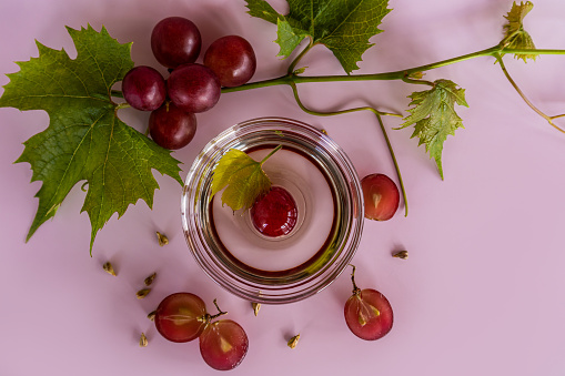 glass bowl with organic grape seed oil, ripe red grape berries. top view. lilac background. high content of vitamins