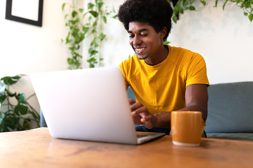 Happy young African American man sitting on couch using laptop to work at home. Working from home concept.