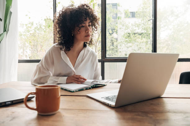 istock Young pretty mixed race woman studying at home with laptop. Taking notes in notebook. Bright spacious living room. 1434378354