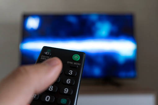 Close-up of a Hand Holding a Remote Control, pointing towards TV.