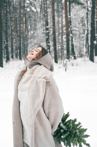 Young beautiful girl in snowy winter forest with christmas tree. Walking, having fun, laughing in stylish clothes, fur coat, woolen shawl, bonnet. Snow lovestory. Romantic date, weekend.