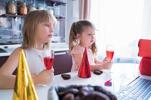 Kids virtual birthday party.Girls in dresses and caps celebrate holiday with friends on isolation.Online conference,video call in laptop,computer.Animator,juice,cookies.Quarantine,coronavirus covid-19