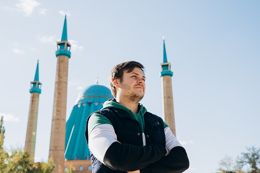 Looking away young man 34 years old against Muslim Mosque (Mashkhur Jusup Central Mosque) and autumn leaves trees. Russian migrant in Kazakhstan. Waist up lifestyle travel portrait, religion concept.