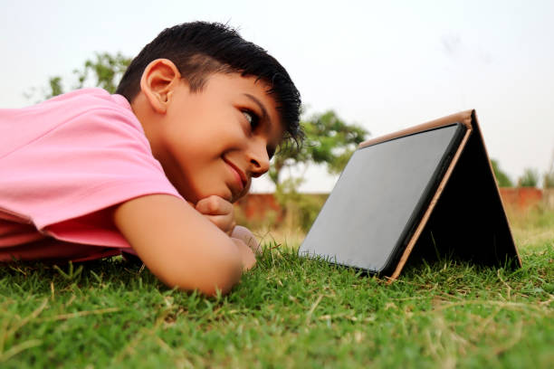 Elementary age child of Indian ethnicity lying down in the grass outdoor in nature during springtime and using Digital Tablet portrait close up.