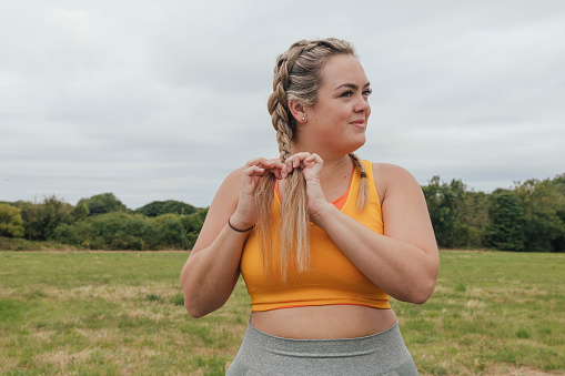 A front-view waist up shot of a young woman standing outdoors on a field in Northumberland, England. She is tying her hair and preparing for a run.