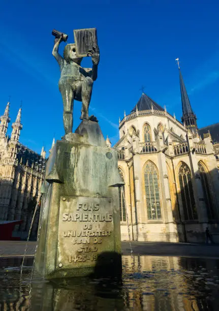 View of Fonske statue and fountain on central square of Leuven city on background of impressive gothic buildings of Town hall and Saint Peter Church on sunny day, Belgium