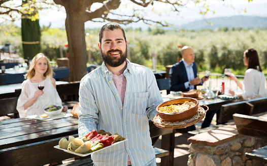 Polite bearded waiter with serving tray welcoming on terrace of country restaurant