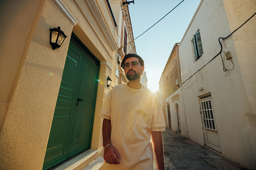 A young and handsome Caucasian man is walking down a typical narrow Mediterranean street. He is very relaxed, elegant and stylish.