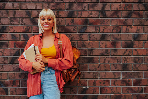 A young female Caucasian student with casual clothes holding books and standing in front of a brick wall at the university with copy space.