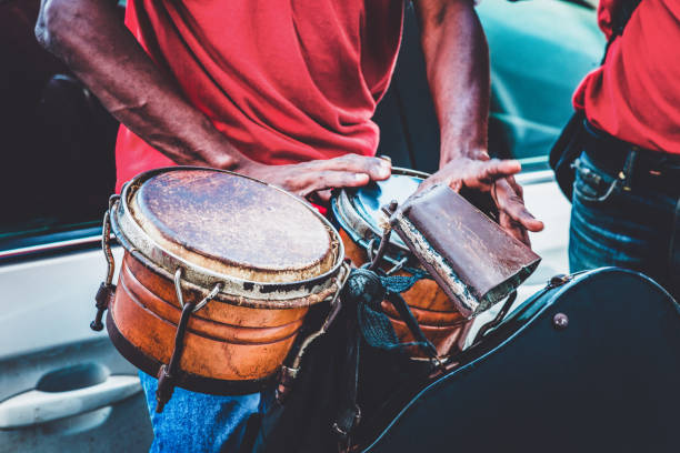 Male musician smiles slightly while playing conga drums in a studio setting. Captured with a dark background and natural expression.