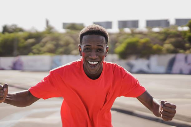 young african man football supporter fan watching soccer match event at stadium - person with red t-shirts having excited fun on sport world championship concept - england football shirt 2022 個照片及圖片檔