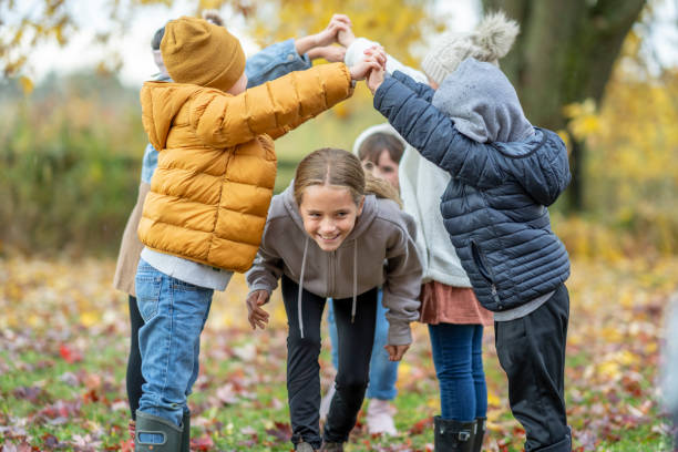 A small group of school-aged children play a game of London Bridges as they hold hands and run under the tunnel of arms.  They are each dressed in warm fall layers and they enjoy the crisp Autumn air.