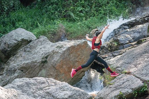 Young woman jumping high over rocky trail while running in green forest in mountains