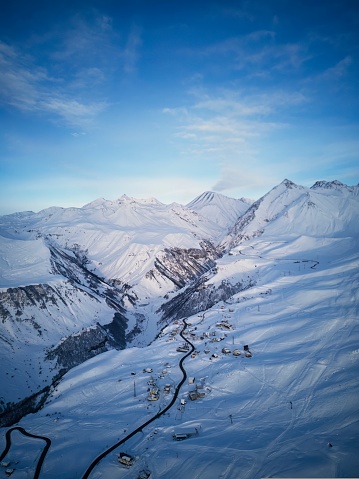 Aerial of snowy mountain range on winter sunrise at ski resort. Drone above mountains valley and village with curvy road at sunset. Caucasus peaks skyline in a pink twilight afterglow.