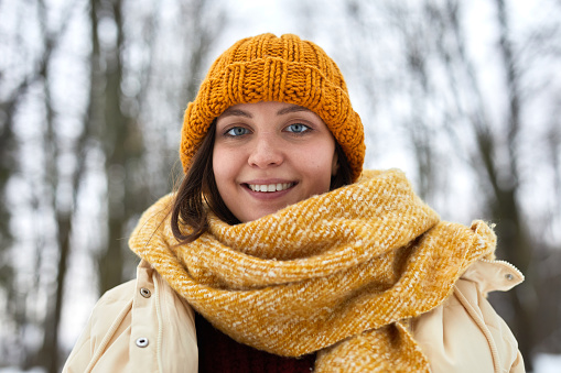 Front view portrait of Caucasian young woman wearing knit hat and scarf in winter forest and smiling at camera