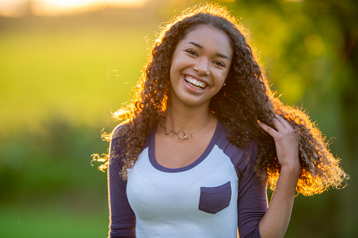 A young female teen of African decent, stands outside on a sunny evening as she poses for a portrait.  She is dressed casually and smiling.