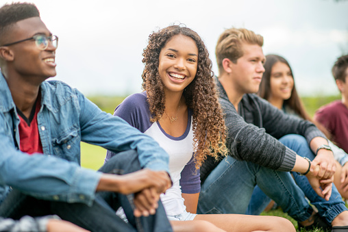 A small group of teenagers sit in the grass as they hang out talking and enjoying a few laughs together.  They are each dressed casually and are smiling as they enjoy the fresh air.