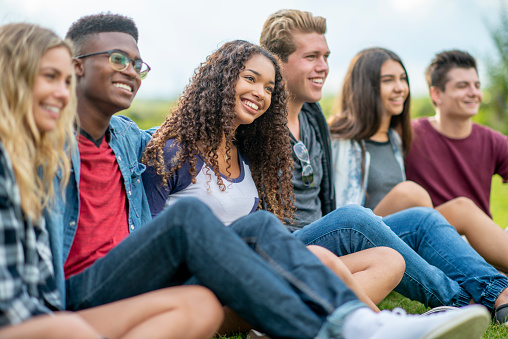 A small group of teenagers sit in the grass as they hang out talking and enjoying a few laughs together.  They are each dressed casually and are smiling as they enjoy the fresh air.