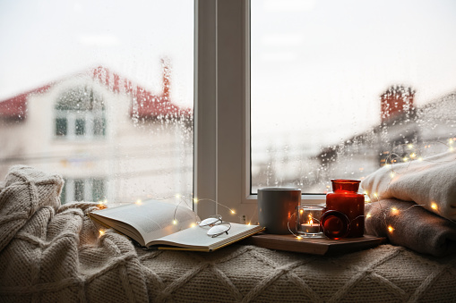 Cup of hot drink, open book and Christmas lights on knitted blanket near window. Cozy season