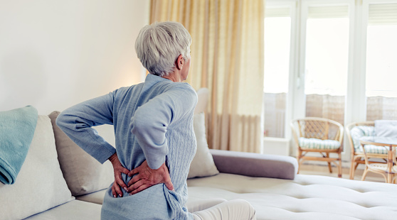 Tired upset elderly woman feeling strong lower back pain after sleeping in incorrect posture on uncomfortable mattress. Senior 60s lady with closed eyes sitting on bed, holding painful lumber muscles.