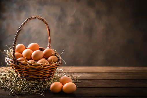 Front view of a wicker basket full of chicken eggs on a rustic wooden table. There are three eggs on the table.