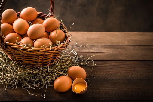 High angle view of a wicker basket full of chicken eggs on a rustic wooden table. There are three eggs on the table and one of them is broken so the egg yolk can be seen.