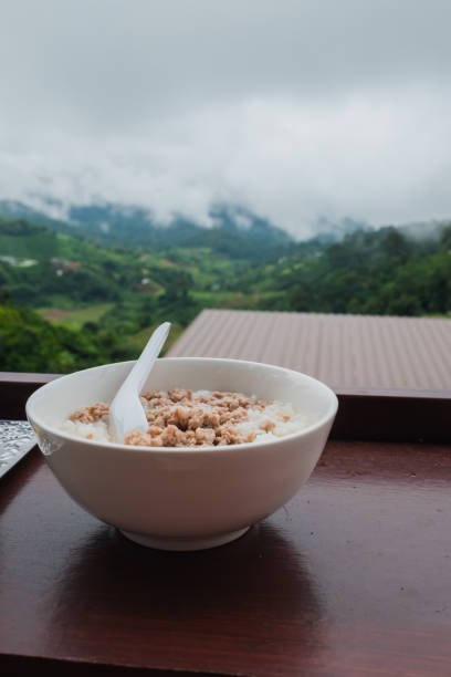 Side portrait of a plate of white pork porridge for breakfast placed on a brown table. Blurred background of mountain view on vacation stock photo