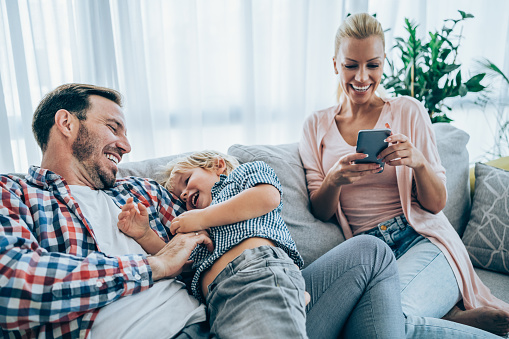Shot of a father and his little boy spending time together at home. Happy father and son having fun in the living room. Mother sitting next to them and using smartphone.
