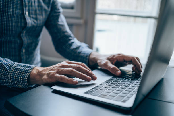 Successful businessman in modern office working on laptop. Cropped shot of a young unrecognizable businessman sitting on desk in modern office and working on laptop. Elegant handsome businessman sitting in his office and using laptop. sales executive stock pictures, royalty-free photos & images