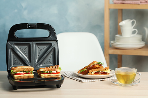 Modern grill maker with sandwiches and tea on light wooden table indoors