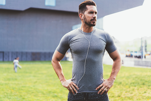 Confident muscular man with hands on waist in sportswear with earphones listening to music preparing for sport exercise standing in street looking away