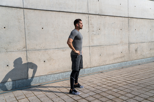 Side view of active muscular adult male in sportswear with earphones standing next to concrete wall and enjoying sunny day while relaxing after training in city