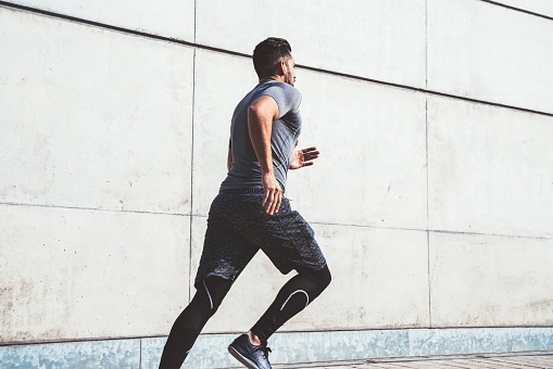 From below of strong guy in sport clothes doing workout exercise training legs running in street of modern city on sunny day