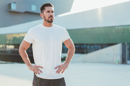 Serious caucasian hipster guy in white shirt with copy space area for brand name or label prepare for morning workout, confident bearded man in earphones standing on stadium ready to exercising
