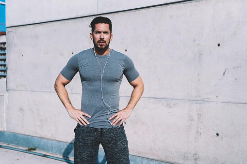 Half length portrait of handsome muscular male in active wear listening music in earphones and resting on break, young confident sportsman prepare for workout keeping perfect body shape and wellness