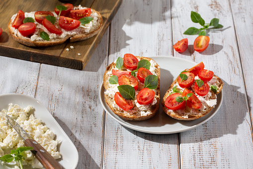 Sandwich with cottage cheese, tomatoes and basil on white wooden background. Traditional Italian bruschetta. Healthy savory feta and tomato toast. Close-up.