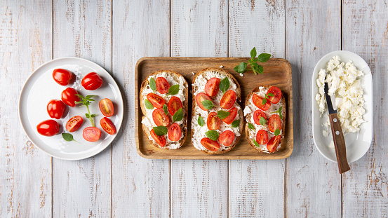 Sandwich with cottage cheese, tomatoes and basil on white wooden background. Traditional Italian bruschetta. Healthy savory feta and tomato toast. Top view.