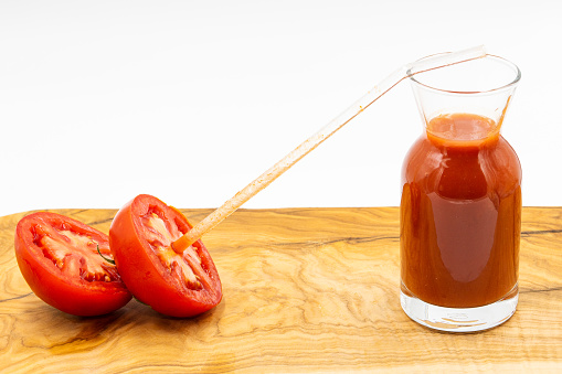 Glass with tomato juice on wooden serving plate and glass straw inside a cut tomato on white background.
