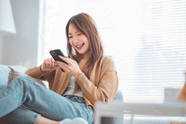 istock Happy young asian woman browsing surfing wireless internet on mobile phone while sitting a couch in living room at home. 1434454974